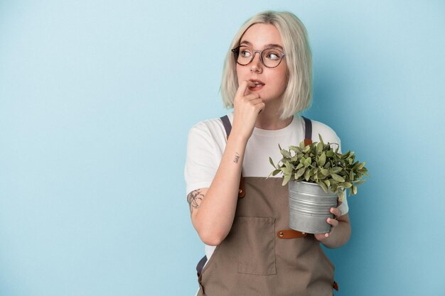 Una joven jardinera caucásica sosteniendo una planta aislada de fondo azul se relajó pensando en algo mirando un espacio de copia.