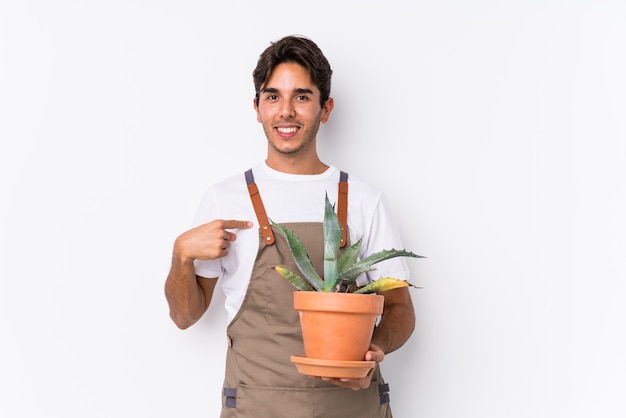 Joven jardinero caucásico hombre sosteniendo una planta aislada persona apuntando con la mano a una camisa copia espacio, orgulloso y confiado