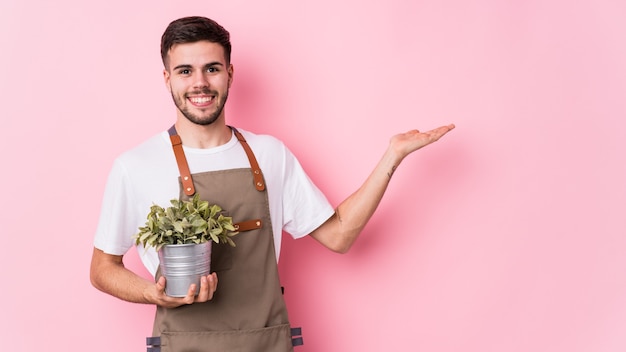 Joven jardinero caucásico sosteniendo una planta aislada mostrando un espacio de copia en una palma y sosteniendo otra mano en la cintura.