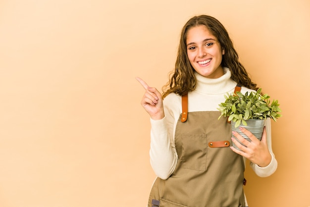 Joven jardinero caucásico sosteniendo una planta aislada sonriendo y apuntando a un lado, mostrando algo en el espacio en blanco.