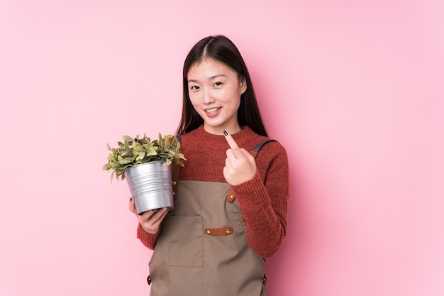 Joven jardinero chino sosteniendo una planta aislada apuntando con el dedo hacia usted como si estuviera invitando a acercarse.