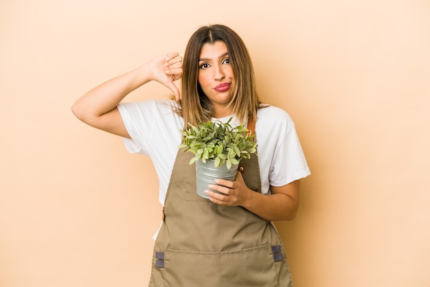 Joven jardinero indio sosteniendo una planta aislada mostrando un gesto de aversión, pulgares hacia abajo. Concepto de desacuerdo.