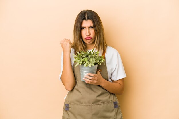 Joven jardinero indio sosteniendo una planta aislada mostrando el puño a la cámara, expresión facial agresiva.