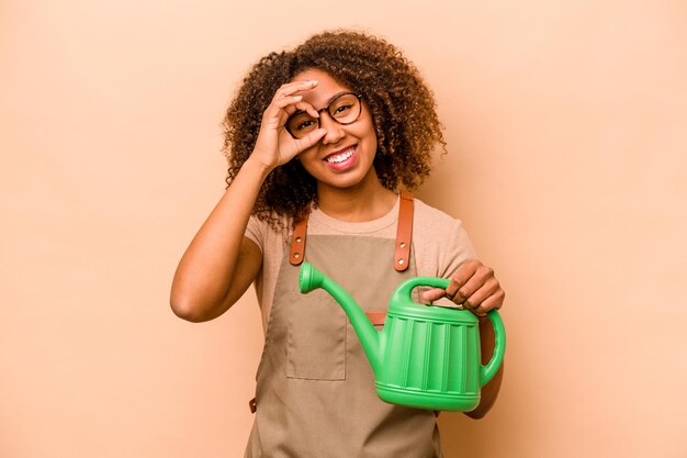 Joven jardinero mujer afroamericana sosteniendo riego aislado sobre fondo beige emocionado manteniendo el gesto de ok en el ojo