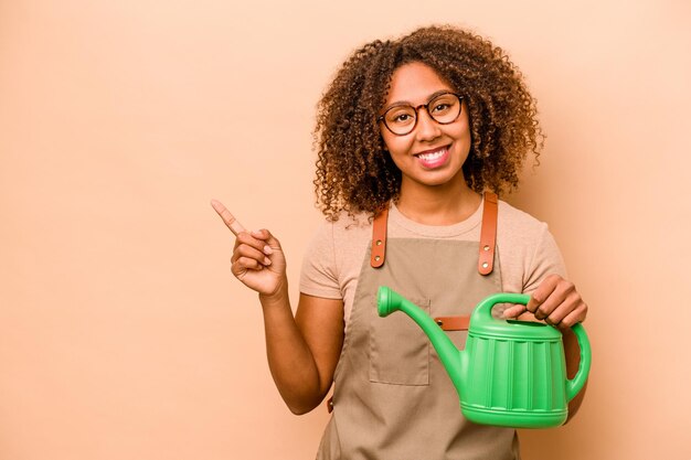 Joven jardinero mujer afroamericana sosteniendo riego aislado sobre fondo beige sonriendo y señalando a un lado mostrando algo en el espacio en blanco