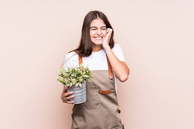 Joven jardinero mujer caucásica sosteniendo una planta aislada cubriendo las orejas con las manos.