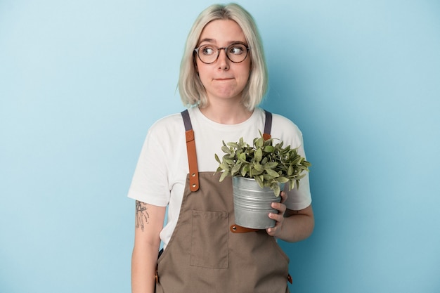 Joven jardinero mujer caucásica sosteniendo una planta aislada sobre fondo azul confundida, se siente dudosa e insegura.