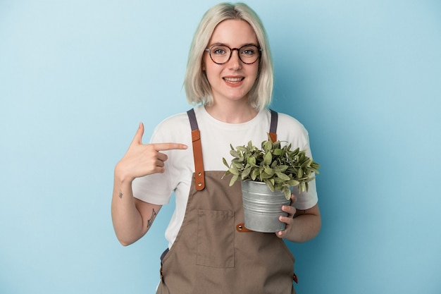 Joven jardinero mujer caucásica sosteniendo una planta aislada sobre fondo azul persona apuntando con la mano a un espacio de copia de camisa, orgulloso y seguro