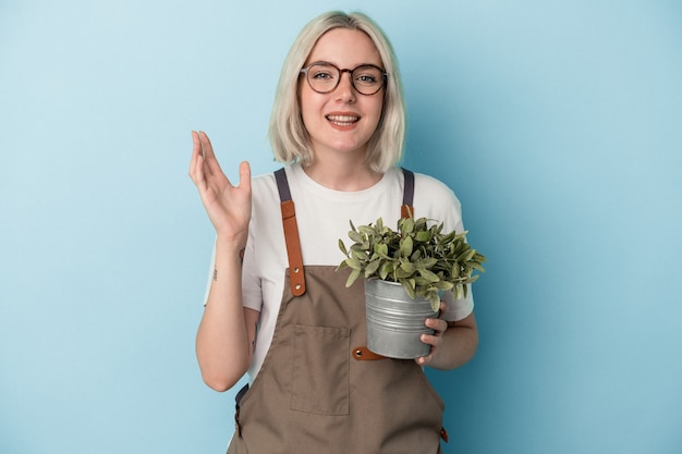 Joven jardinero mujer caucásica sosteniendo una planta aislada sobre fondo azul recibiendo una agradable sorpresa, emocionada y levantando las manos.