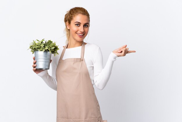 Joven jardinero rubio mujer chica sosteniendo una planta sobre pared blanca aislada que señala con el dedo hacia el lado