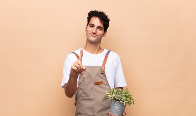 Joven jardinero sonriendo con orgullo y confianza haciendo la pose número uno triunfalmente, sintiéndose como un líder