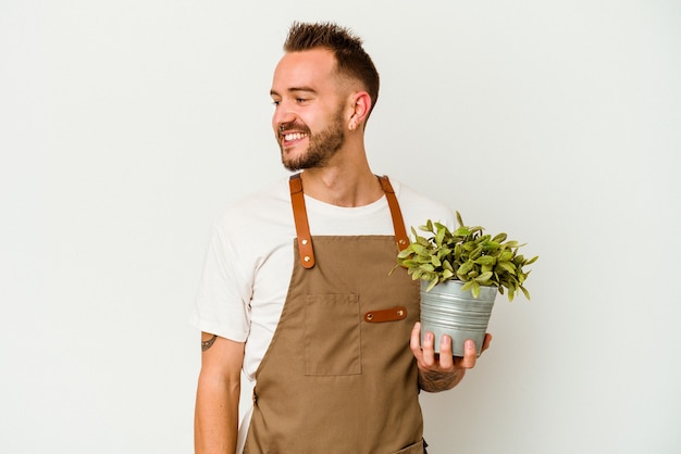 Joven jardinero tatuado caucásico sosteniendo una planta aislada sobre fondo blanco mira a un lado sonriente, alegre y agradable.