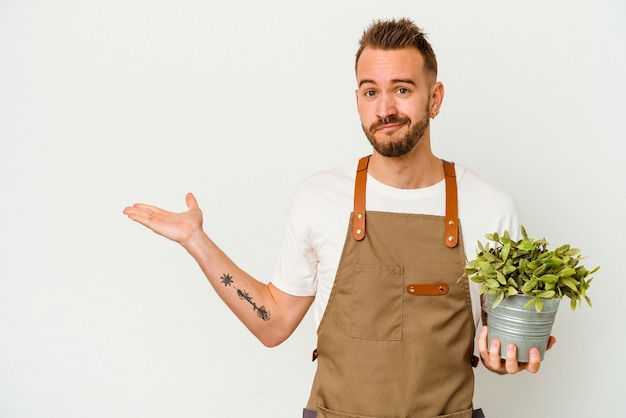 Joven jardinero tatuado hombre caucásico sosteniendo una planta aislada sobre fondo blanco mostrando un espacio de copia en una palma y sosteniendo otra mano en la cintura.