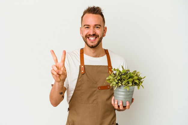 Joven jardinero tatuado hombre caucásico sosteniendo una planta aislada sobre fondo blanco mostrando el número dos con los dedos.