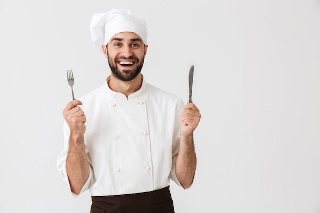 Joven jefe en uniforme de cocinero sonriendo mientras sostiene una cuchara y un tenedor de metal aislado sobre la pared blanca