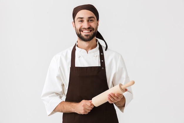 Joven jefe en uniforme de cocinero sonriendo y sosteniendo un rodillo de madera de cocina aislado sobre una pared blanca