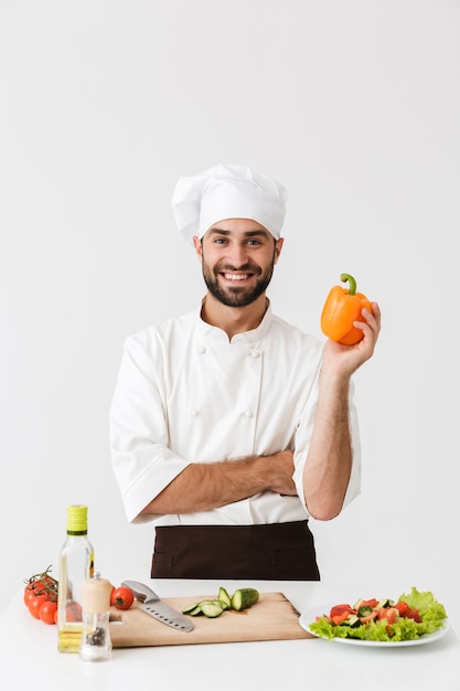 Joven jefe en uniforme sosteniendo papel dulce mientras cocina ensalada de verduras sobre tabla de cortar de madera aislada sobre pared blanca