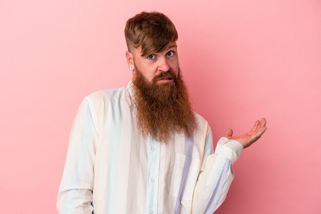 Foto joven jengibre caucásico con barba larga aislado sobre fondo rosa dudando y encogiéndose de hombros en gesto de interrogación.