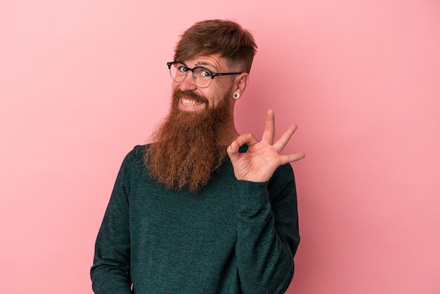 Foto joven jengibre caucásico con barba larga aislado sobre fondo rosa guiña un ojo y tiene un gesto bien con la mano.