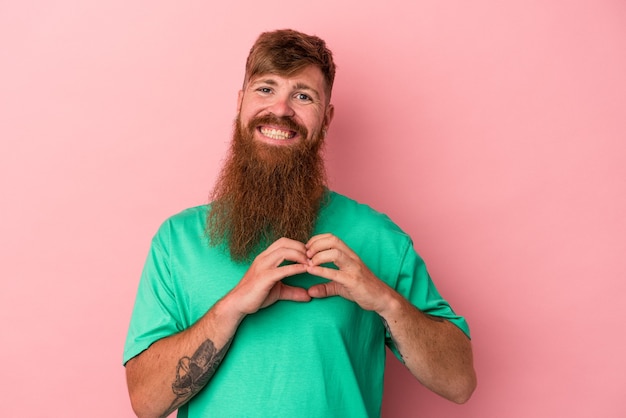 Joven jengibre caucásico con barba larga aislado sobre fondo rosa sonriendo y mostrando una forma de corazón con las manos.