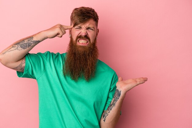 Foto joven jengibre caucásico con barba larga aislado sobre fondo rosa sosteniendo y mostrando un producto en la mano.