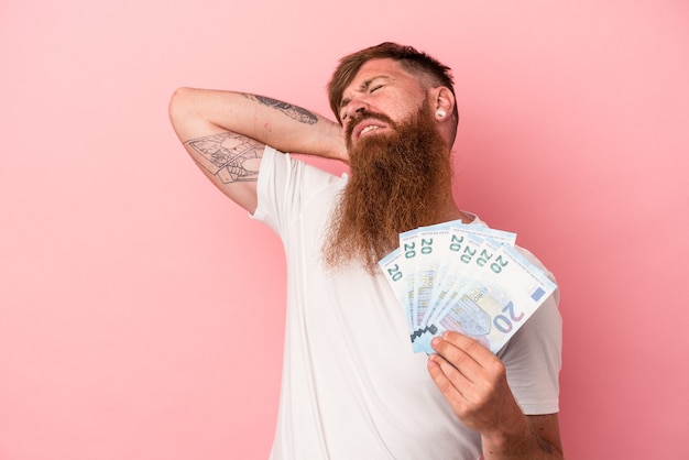 Joven jengibre caucásico con barba larga sosteniendo billetes aislados sobre fondo rosa tocando la parte posterior de la cabeza, pensando y haciendo una elección.