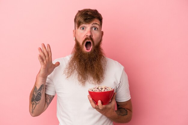 Joven jengibre caucásico con barba larga sosteniendo un cuenco de cereales aislado sobre fondo rosa sorprendido y consternado.