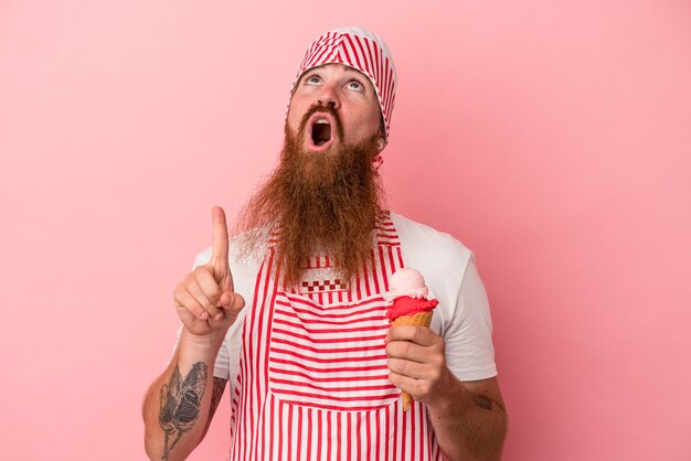 Joven jengibre caucásico con barba larga sosteniendo un helado aislado sobre fondo rosa apuntando hacia arriba con la boca abierta.