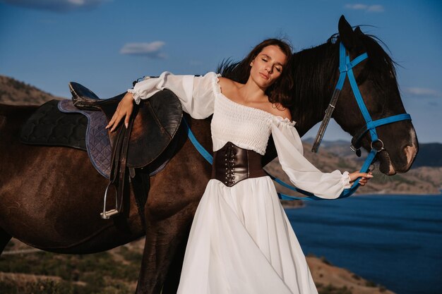 Una joven jinete posa junto a su caballo en el contexto de un paisaje de montaña y mar. El concepto de montar a caballo. Fotografía artística. Portada confeccionada para libros y revistas.