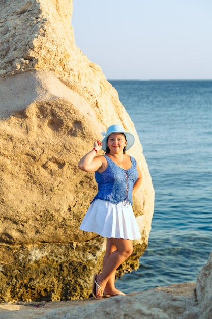 Una joven judía con una falda blanca y un sombrero azul en la orilla del mar cerca de las rocas Vertical