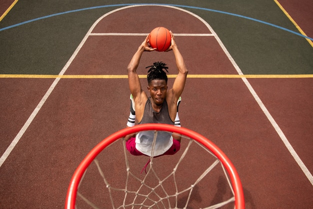 Joven jugador de baloncesto africano haciendo esfuerzo mientras salta y lanza la pelota en la canasta durante el entrenamiento