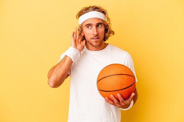 Joven jugador de baloncesto hombre caucásico aislado de fondo amarillo tratando de escuchar un chisme.