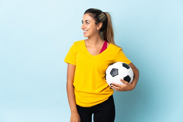 Joven jugador de fútbol hispano mujer sobre aislado en la pared azul mirando hacia el lado