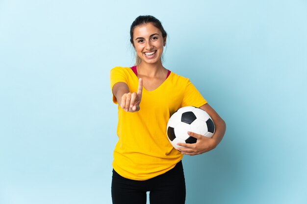 Joven jugador de fútbol hispano mujer sobre aislado en la pared azul mostrando y levantando un dedo