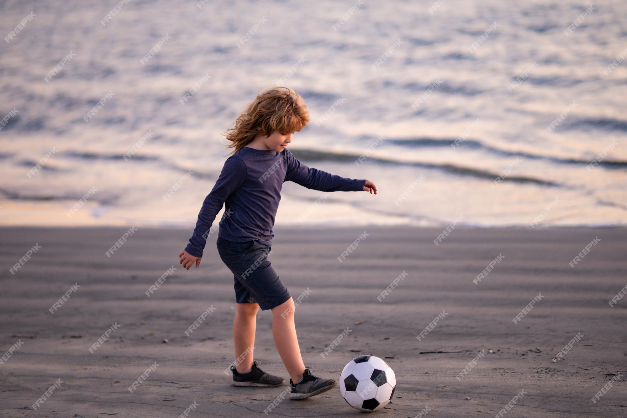 de fútbol en ropa deportiva con de fútbol un niño alegre disfruta de los juegos de fútbol en la playa al atardecer | Foto Premium