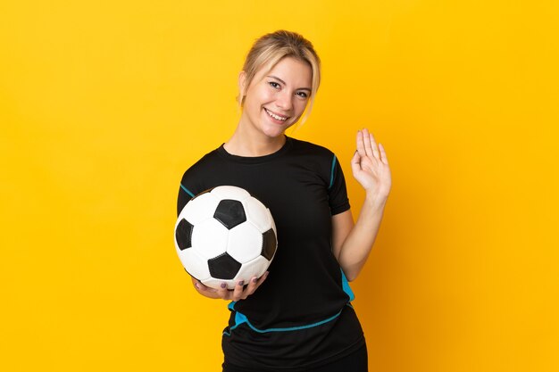 Joven jugador de fútbol ruso mujer aislada sobre fondo amarillo saludando con la mano con expresión feliz