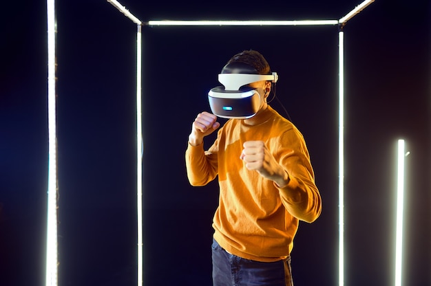 Joven jugador luchando en casco de realidad virtual y gamepad en cubo luminoso, vista frontal. Interior oscuro del club de juego, foco en el fondo, tecnología VR con visión 3D