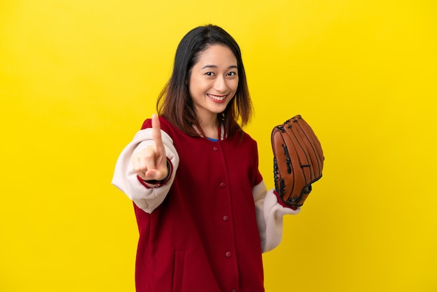 Joven jugadora vietnamita con guante de béisbol aislada de fondo amarillo mostrando y levantando un dedo
