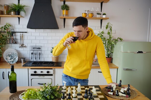 Joven jugando al ajedrez en la mesa de la cocina y bebiendo vino tinto Hombre con alcohol juega en un juego de mesa lógico con él mismo parado en la cocina