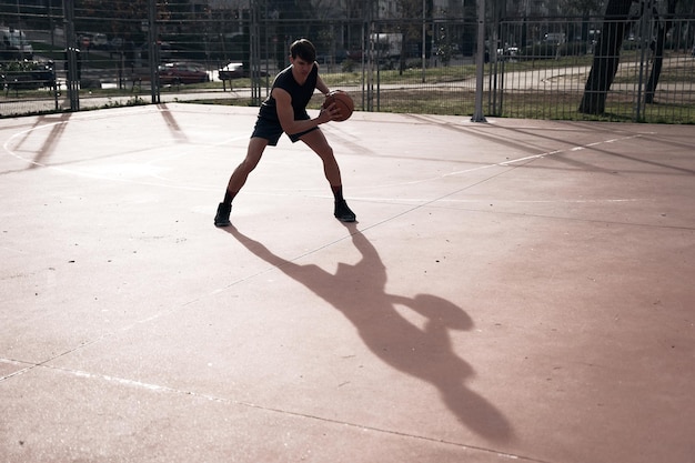 Joven jugando baloncesto al aire libre en la calle con largas sombras