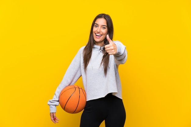 Joven jugando baloncesto sobre una pared amarilla aislada con los pulgares arriba porque algo bueno ha sucedido