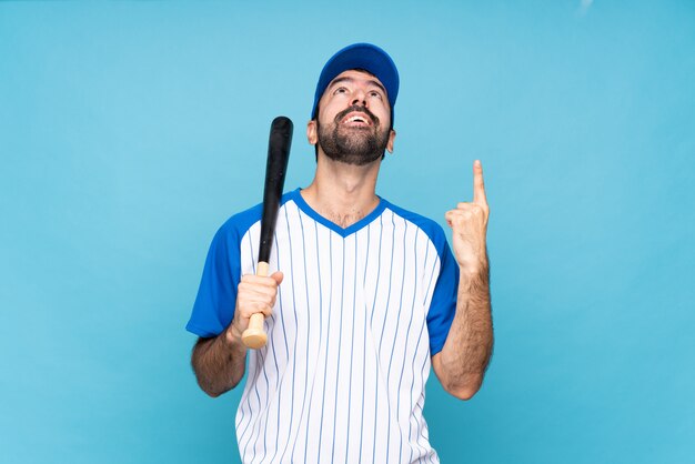 Joven jugando béisbol sobre pared azul aislado apuntando hacia arriba y sorprendido