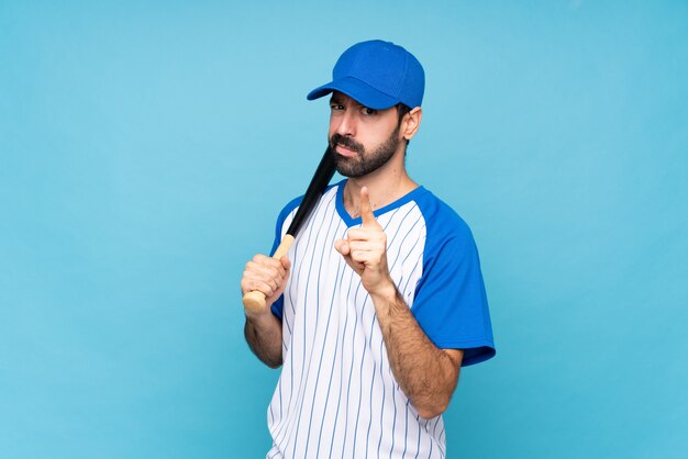 Joven jugando béisbol sobre pared azul aislado frustrado y apuntando hacia el frente