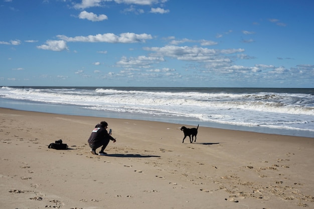 Joven jugando con perro negro en la playa bajo un cielo azul con nubes.