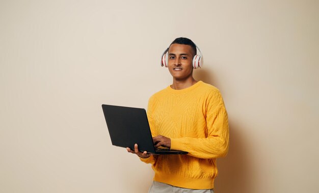 Joven latín con auriculares sosteniendo una computadora portátil sonriendo mirando a la cámara sobre un fondo marrón