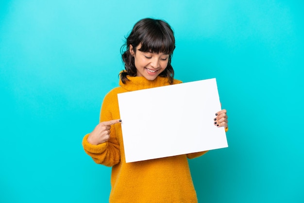 Joven latina aislada de fondo azul sosteniendo un cartel vacío con expresión feliz y apuntándolo