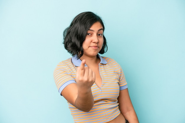 Joven latina aislada sobre fondo azul apuntando con el dedo como si invitara a acercarse.