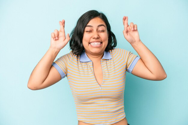 Joven latina aislada sobre fondo azul cruzando los dedos para tener suerte