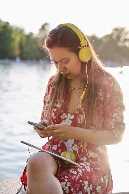 Joven latina con auriculares sentada en un parque charlando en su teléfono inteligente