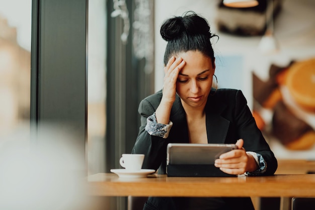 Joven latina con cara triste sentada en la cafetería en una mesa de madera, tomando café y usando una laptop.